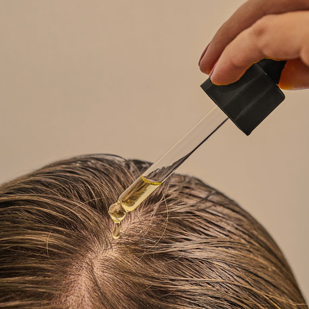 Hand using a dropper to apply oil to hair near the scalp against a neutral background