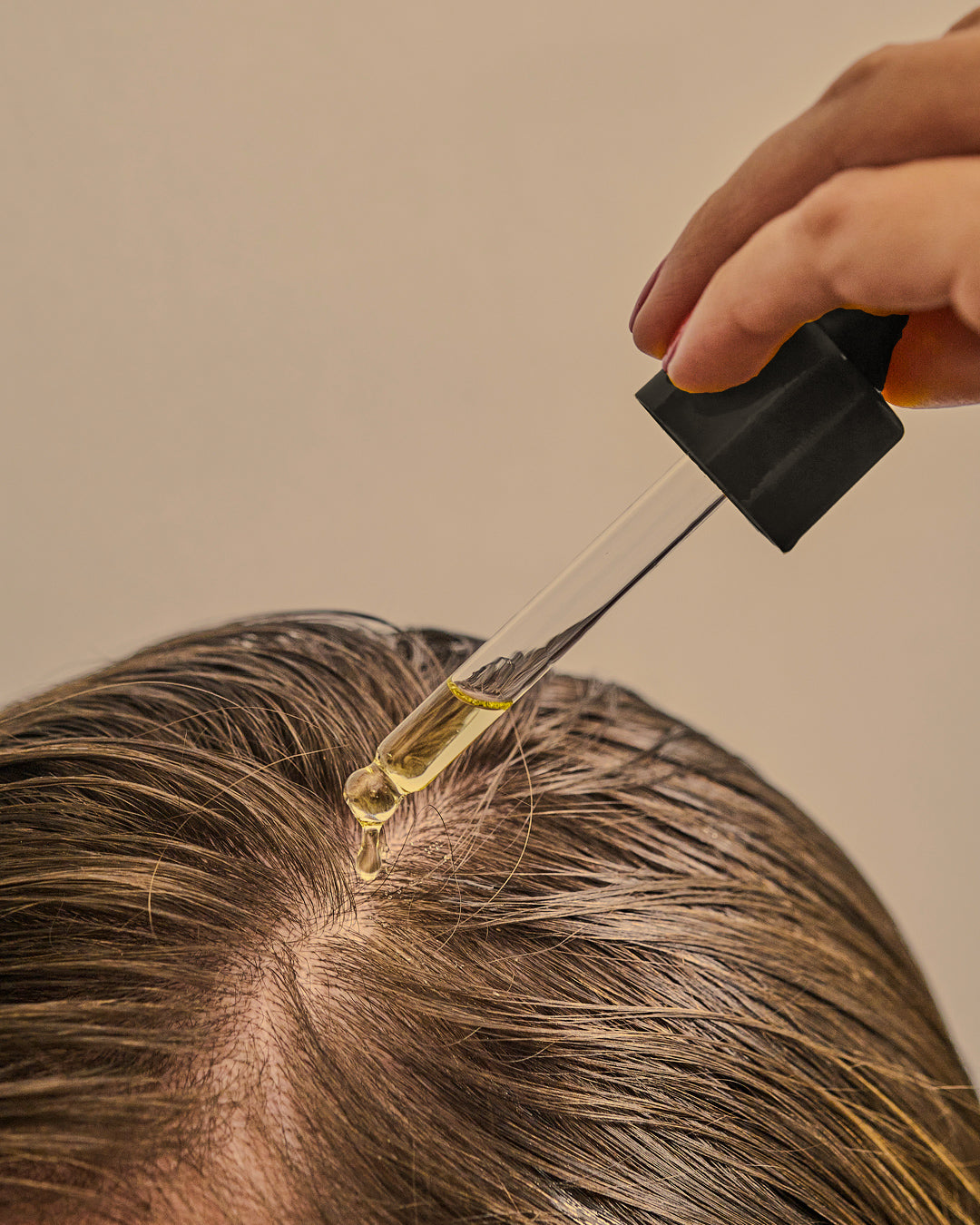 Hand using a dropper to apply oil to hair near the scalp against a neutral background