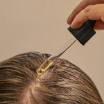 Hand using a dropper to apply oil to hair near the scalp against a neutral background